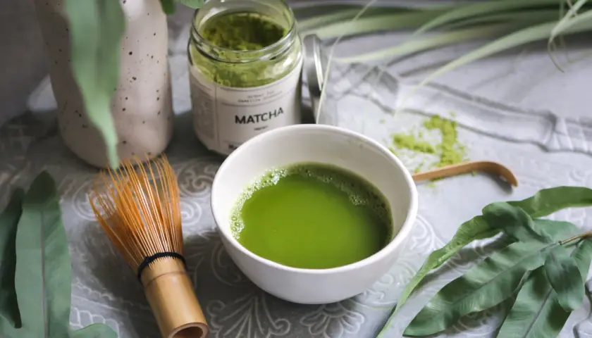 Matcha-Pulver in einem Glas, eine Schale mit zubereitetem Matcha und ein Bambusbesen. Die Matcha-Wirkung auf den Darm wird oft positiv beschrieben.