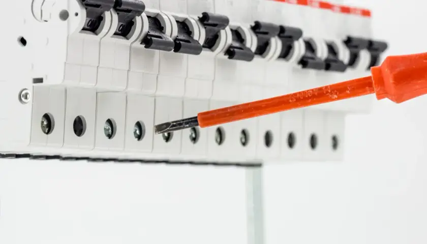 Elektryk dokręca śrubę w skrzynce z bezpiecznikami, przygotowując instalację. Widać przewód uziemienia i narzędzie rcd.