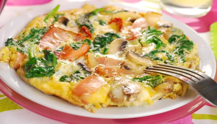 Omlet z szpinakiem, pieczarkami i bekonem na białym talerzu, widelcem.