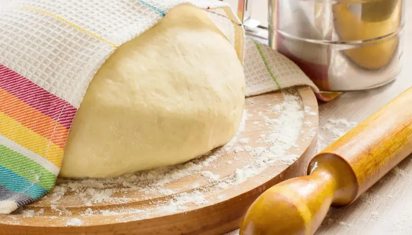 Masa de pan cubierta con un paño a rayas, sobre una tabla de madera con harina esparcida. Rodillo y tamizador de harina, ingredientes para hacer pan.