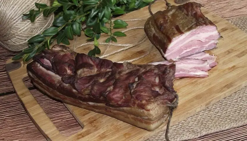 Dwa kawałki wędzonego boczku, jeden pokrojony w plastry, leżą na desce. Obok gałązka zieleni i sznurek.