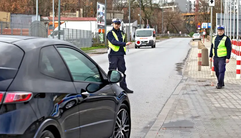 Policjant zatrzymuje czarne auto, być może za recydywę mandatu. Na drodze stoi biały van.