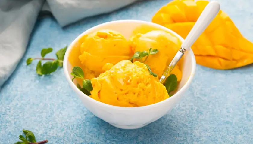 Coppa di sorbetto al mango con menta fresca e una fetta di mango sullo sfondo.