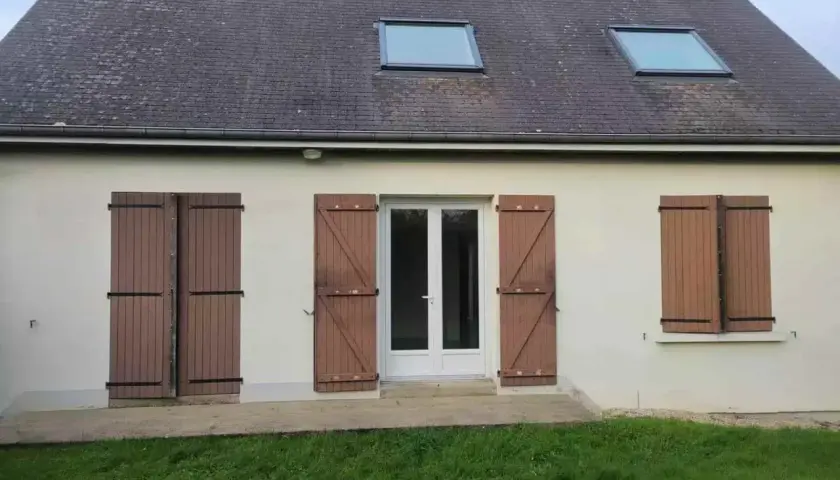 Façade d'une maison année 80, caractéristiques : toit en ardoise, deux fenêtres de toit, porte-fenêtre blanche et volets en bois marron.