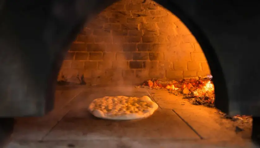 Wnętrze pieca do pizzy z palącym się drewnem i surowym ciastem na kamiennej podłodze. Dym unosi się wokół.
