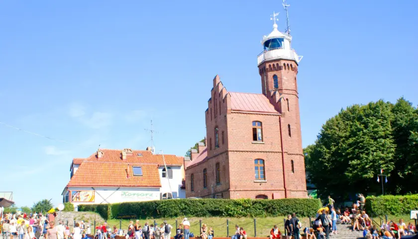 Urokliwa latarnia w Ustce, otoczona zielenią i tłumem turystów.