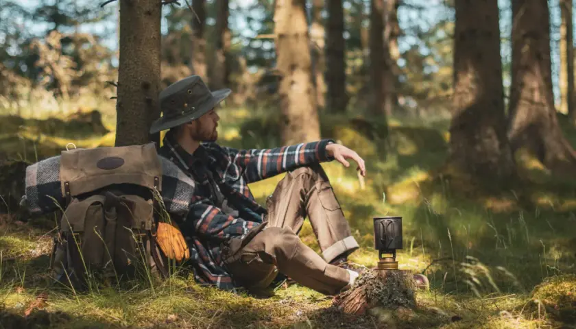 Man med hatt och rutig skjorta vilar vid ett träd under en vandring i Småland. Ryggsäck och stormkök redo för äventyr.