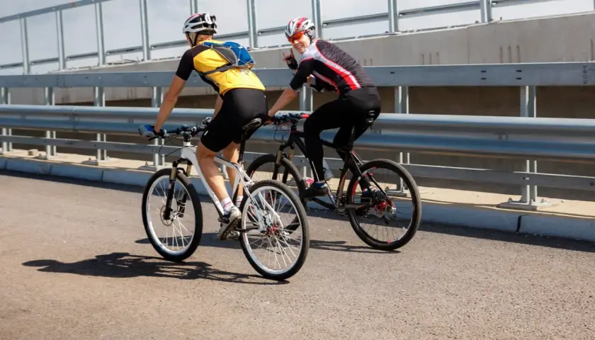 Dwóch rowerzystów jedzie obok siebie po moście. Jeden z nich macha do widza.