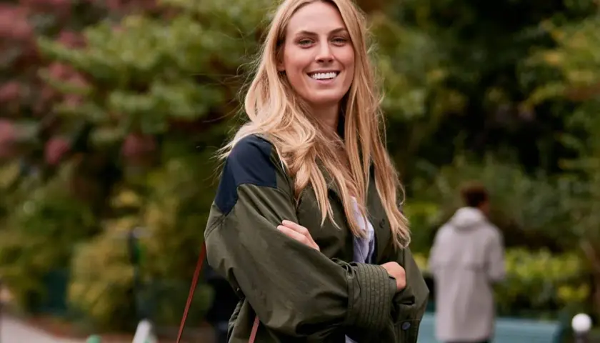 Mujer sonriente con un corte de pelo a capas, luciendo una chaqueta verde y un bolso de cuero.