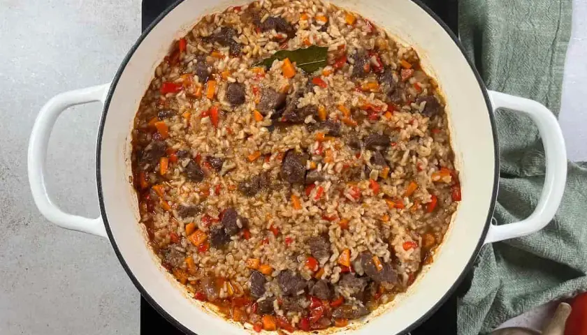Arroz con carne y verduras, cocinado en una olla blanca. Los trozos de carne se mezclan con el arroz y pimientos rojos y zanahorias picadas.