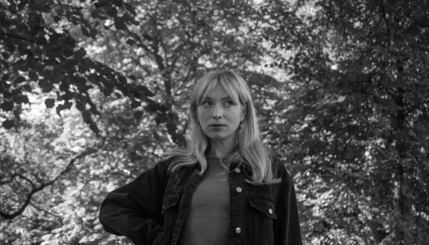 Sandra Beijer, med sitt blonda hår och blick mot horisonten, står i en skog. Hon bär en jeansjacka.