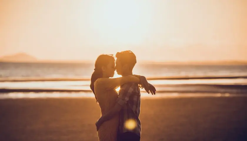 Para zakochanych na plaży o zachodzie słońca. Mężczyzna patrzy kobiecie na usta, obejmując ją.