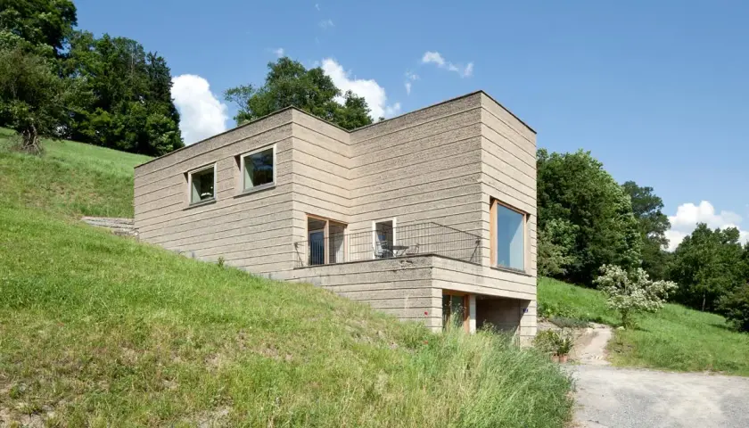 Modernes Haus mit rauer Fassade, das die natürliche Textur von **Lehm als Baustoff** zeigt. Eingebettet in eine grüne Hügellandschaft unter blauem Himmel.