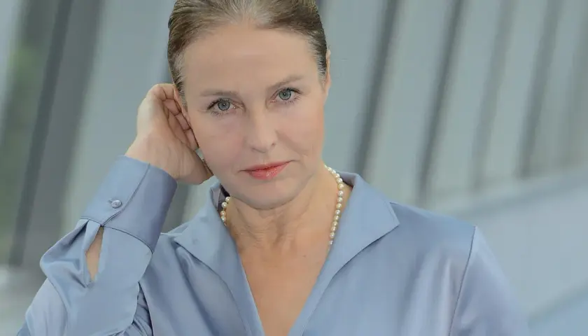 Portret kobiety w niebieskiej koszuli i perłach, opierającej dłoń o ucho.