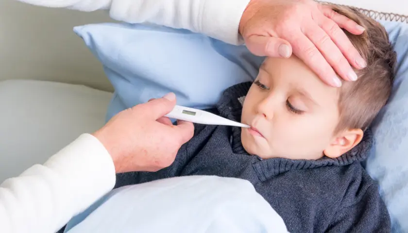 Dziecko leży w łóżku, a opiekunka mierzy mu temperaturę. Dłoń opiekunki delikatnie dotyka czoła malucha, podczas gdy druga trzyma termometr. To obraz troski i dbania o zdrowie, przypominający o tradycyjnych metodach, jak stawianie baniek u dzieci.