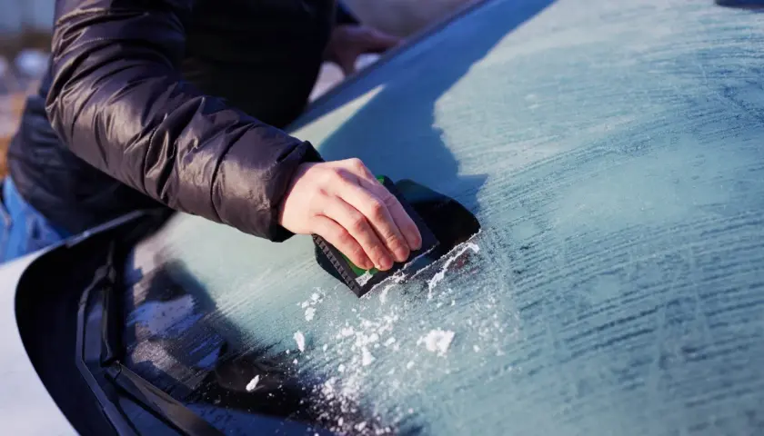 En hand skrapar is från insidan av bilrutan. Snöflingor syns på den frostiga ytan.