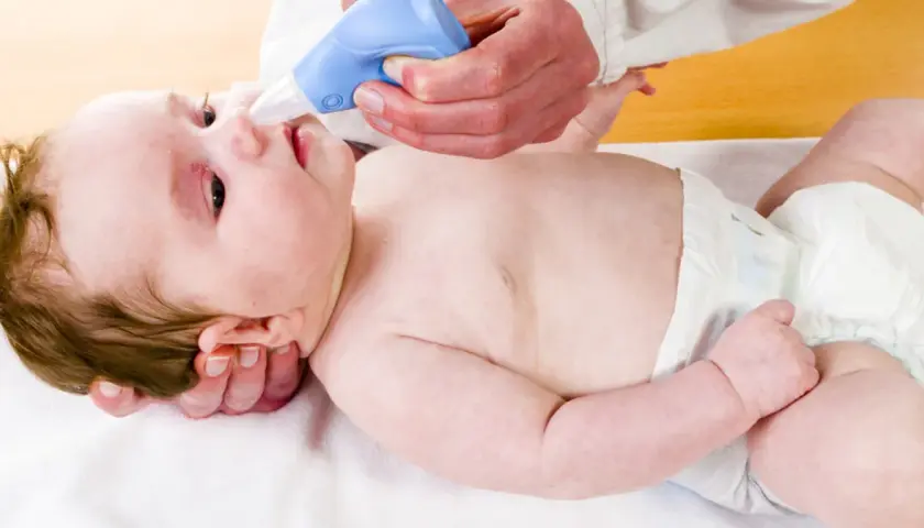 Pomoc w walce z katarem u niemowlaka. Ręce delikatnie trzymają dziecko, a niebieski aspirator do nosa jest używany do oczyszczania jego noska.