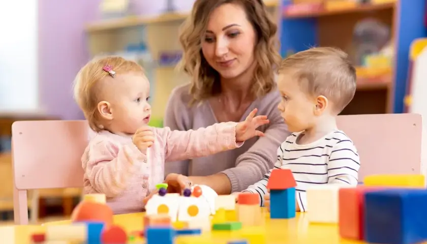 Nauczycielka i dwoje dzieci bawią się klockami przy żółtym stole w żłobku.