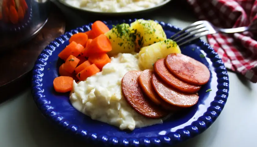 En blå tallrik med stuvad blomkål, kokt potatis, morötter och stekt falukorv. En klassisk svensk middag.