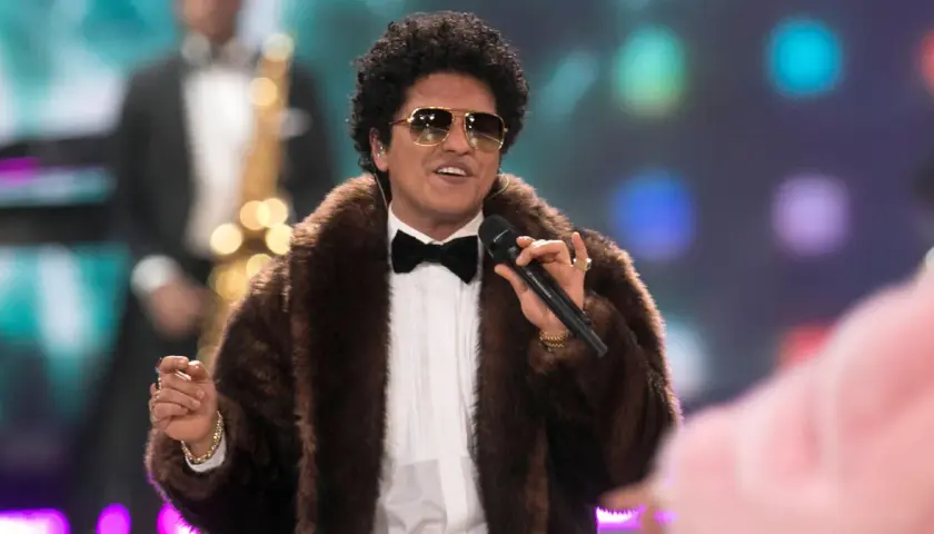 Bruno Mars w futrzanym płaszczu i okularach przeciwsłonecznych śpiewa na koncercie, trzymając mikrofon.