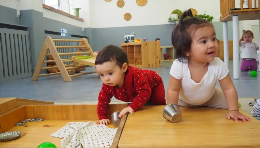 Bebés explorando en una **montessori guarderia**. Un niño gatea con un suéter rojo, mientras otro sonríe.