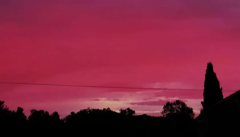 Un ciel rouge le soir, teinté de rose, surplombe une silhouette d'arbres et d'une maison.
