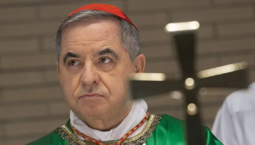 Kardinal Becciu trägt eine rote Pileolus und eine grüne Soutane. Ein Kreuz ist im Vordergrund unscharf.