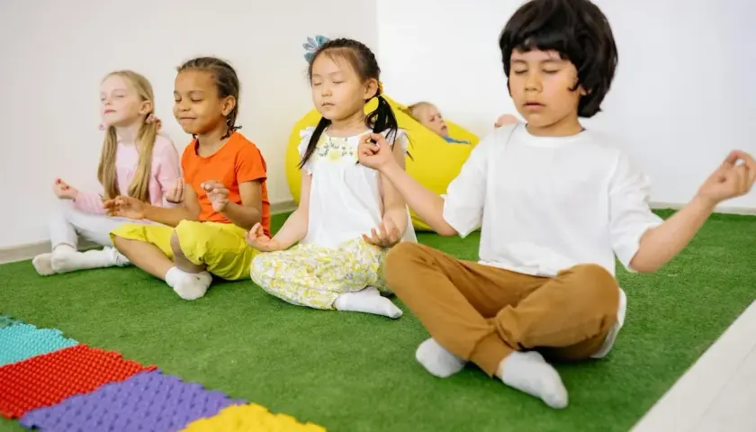 Kinder üben Achtsamkeit im Unterricht, sitzen im Schneidersitz und meditieren. Ein Junge im Vordergrund führt die Übung an.