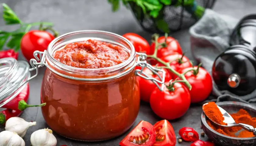 Tarro de cristal lleno de vibrante salsa de tomate casera, rodeado de tomates frescos, ajos, guindillas y especias.
