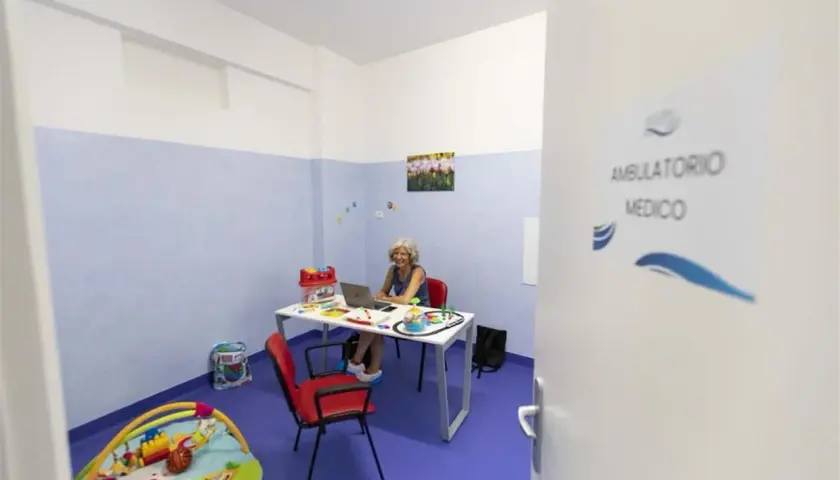 Un ambulatorio medico con una donna al computer, giocattoli e un tappeto gioco. La differenza tra ambulatorio e poliambulatorio è la specializzazione.