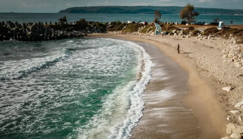 Plaża w Bułgarii, gdzie morze szumi, a fale obmywają brzeg. W oddali widać wzgórza i samochody.