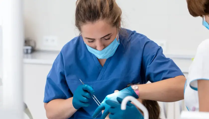 Dentystka w niebieskim fartuchu i masce zakłada opatrunek na zęba pacjentowi.