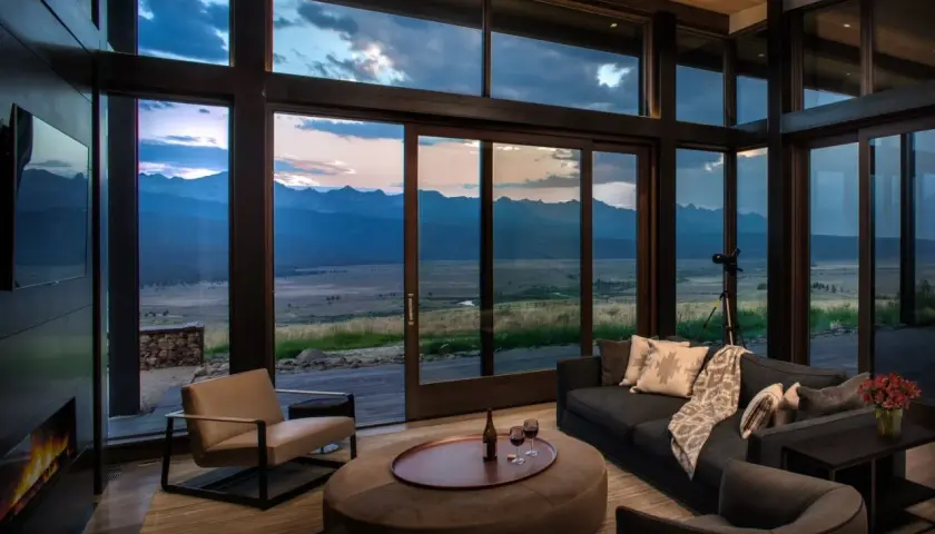 Cozy living room in a modern mountain house, featuring a fireplace, plush seating, and expansive windows with a breathtaking mountain vista at dusk.