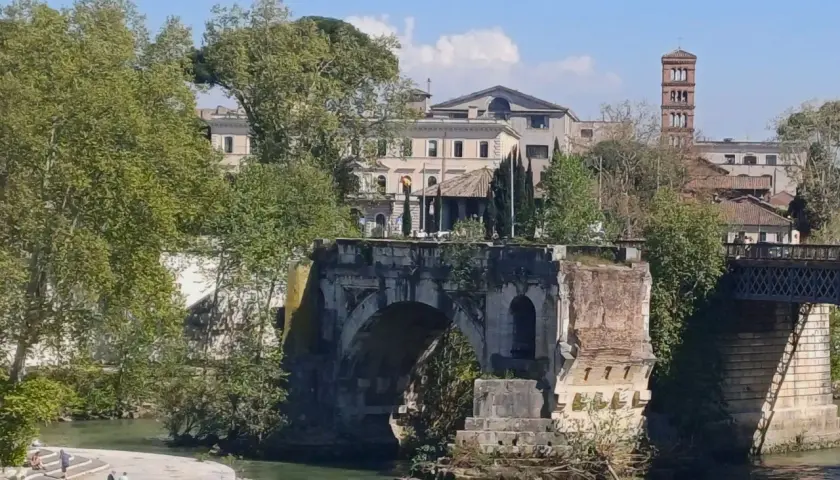 Antico **ponte rotto a Roma**, con rovine e vegetazione che si arrampicano sui resti. In lontananza, edifici storici e una torre campanaria.