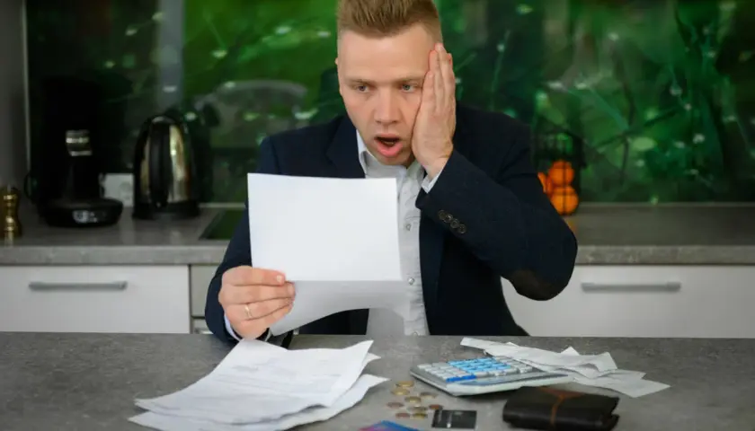 Zaskoczony mężczyzna z papierami i kalkulatorem na stole zastanawia się, jak spłacić debet.