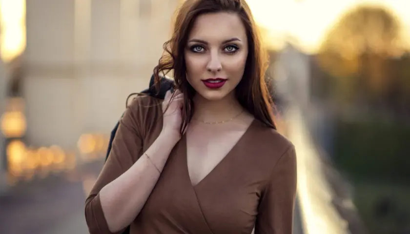 Portret młodej kobiety z burzą brązowych włosów, w brązowej sukience, z kurtką na ramieniu. Ta fotografia ludzi uchwyciła jej spojrzenie.