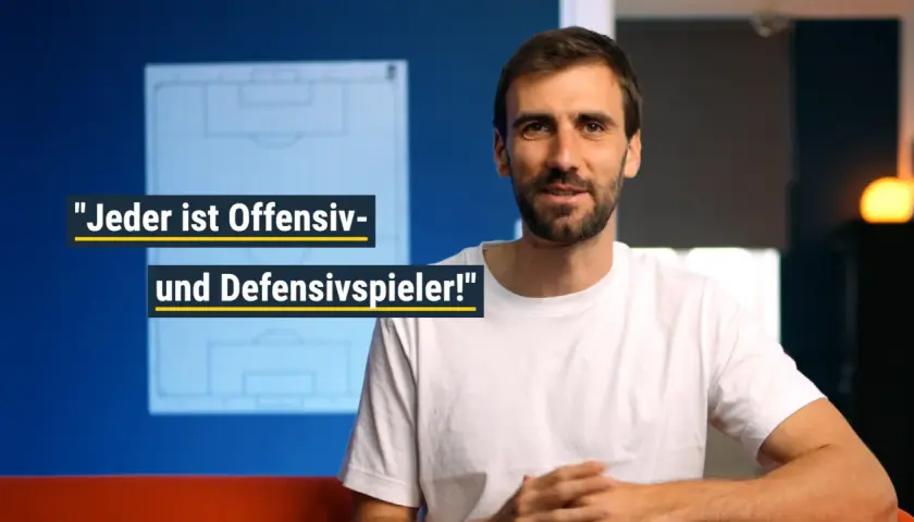 Ein Mann im weißen T-Shirt spricht über Fußballtaktiken. Er erklärt, dass jeder Spieler sowohl offensiv als auch defensiv gegen den Ball spielen muss.