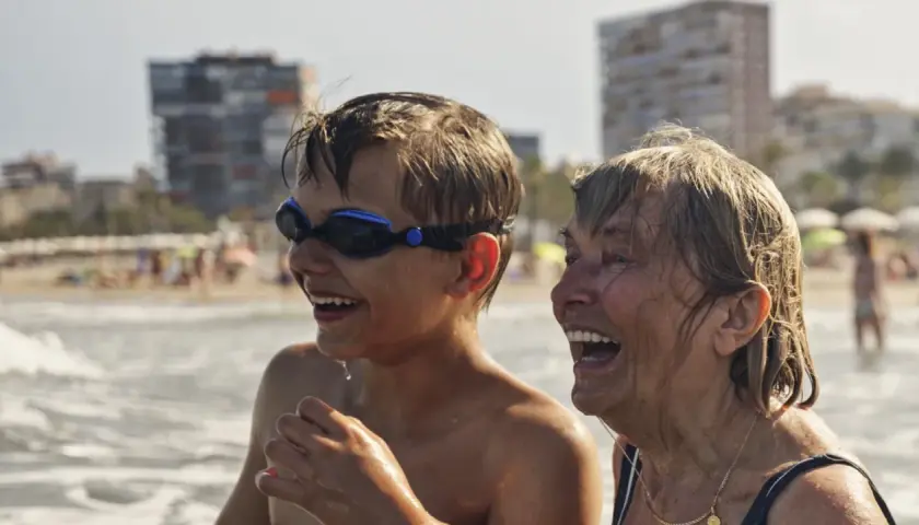 Chłopiec w goglach i starsza kobieta śmieją się na plaży. Wakacje bez zmartwień dzięki najlepszemu ubezpieczeniu turystycznemu.