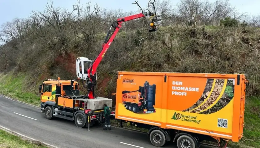 Ein orangefarbener LKW mit einem roten anbaukran schneidet Äste von einem Baum. Daneben steht ein Anhänger mit der Aufschrift "Der Biomasse Profi".
