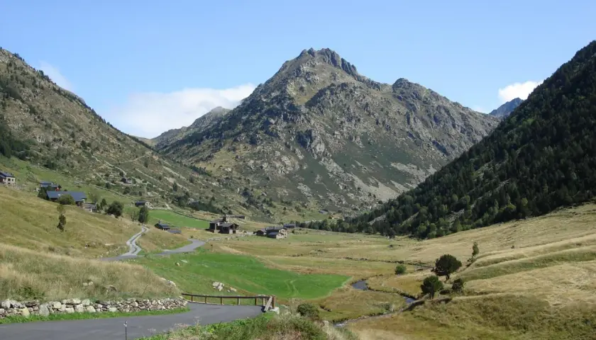 Un camino serpentea hacia el estany de Juclar, un valle andorrano con casas dispersas y un pico imponente.