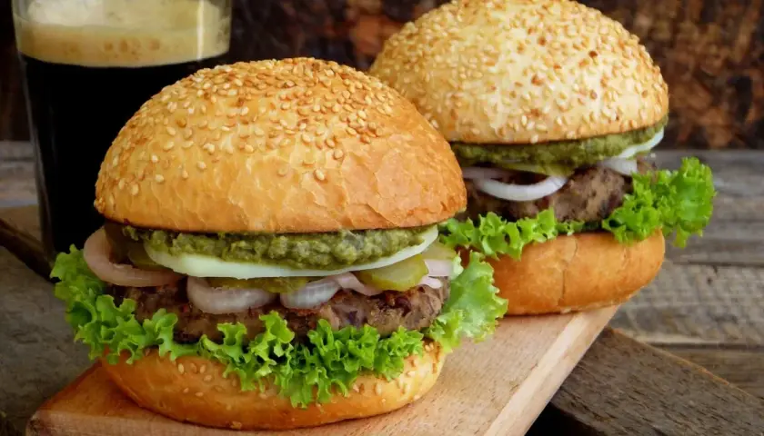 Dwa domowe burgery z sezamem, sałatą, cebulą i sosem, obok szklanki ciemnego napoju.