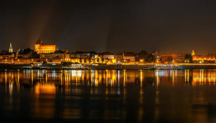 Nocny widok na oświetlony bulwar w Toruniu z odbiciem świateł w Wiśle.