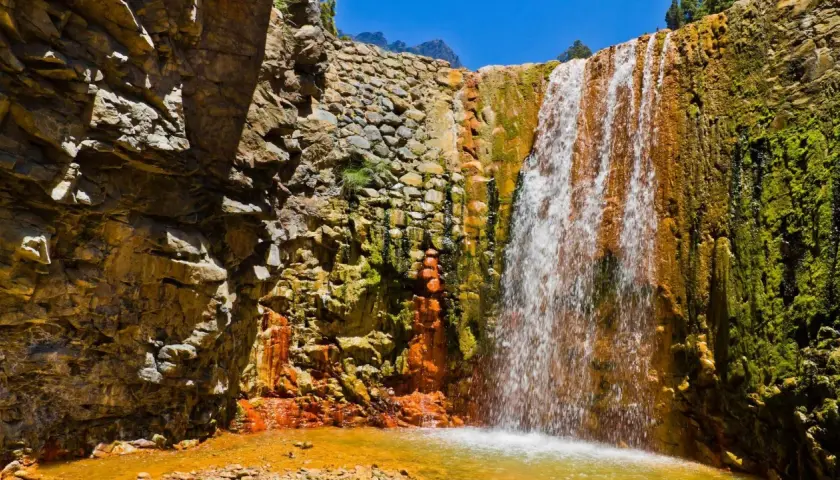 Ein Wasserfall stürzt über moosbewachsene, orangefarbene Felsen. Die Felsen sind von der Sonne beschienen, und das Wasser sammelt sich in einem goldenen Becken. Ein Stück Natur auf La Palma.