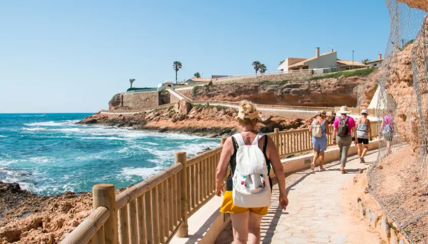 Paseo por la costa en cala capitan. Turistas caminan por un sendero con vistas al mar azul y acantilados rojizos.