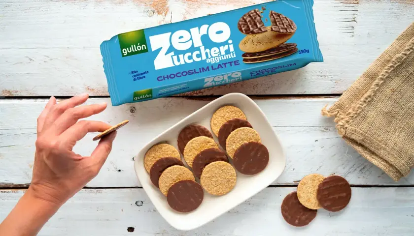 Biscotti senza zucchero Gullón Chocoslim Latte, alcuni ricoperti di cioccolato, su un piatto bianco. Una mano ne prende uno.