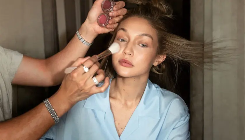 En makeupartist applicerar rouge på en kvinna som håller två små burkar med rouge. Kvinnan har sitt hår uppsatt och bär en ljusblå skjorta.