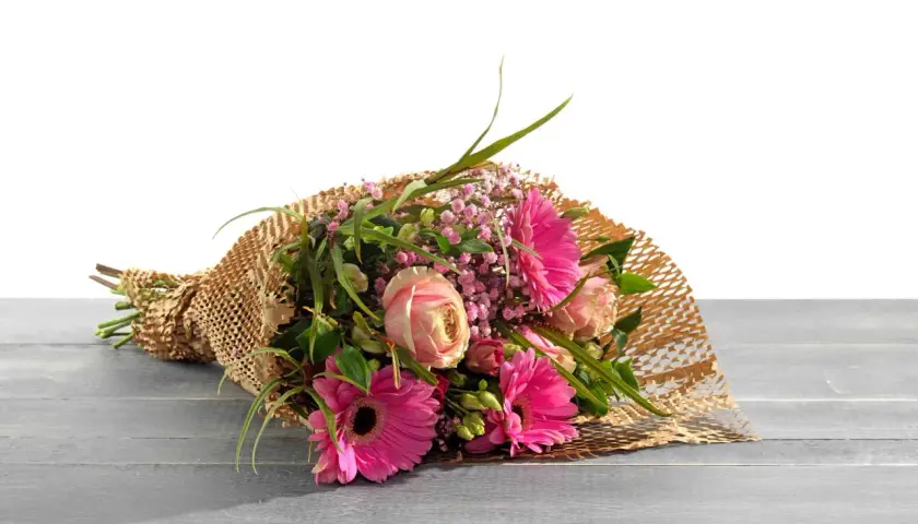 Ein Strauß bunter Blumen, liebevoll als Geschenk verpackt in braunem Papier mit Wabenstruktur, liegt auf einer Holzoberfläche.