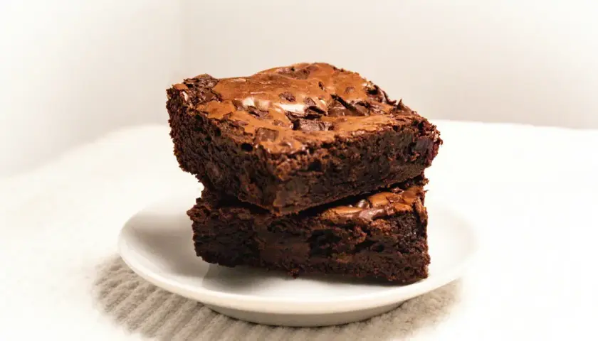 Dwa wilgotne brownie czekoladowe ułożone jedno na drugim na białym talerzyku.