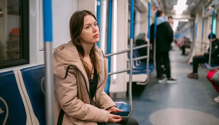 Młoda kobieta w beżowej kurtce siedzi w metrze, trzymając telefon. Jej zamyślona mina sugeruje **wypalenie zawodowe**, być może szuka ulgi lub sposobu na L4.