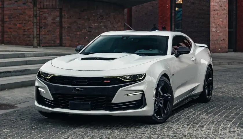 Biały Chevrolet Camaro SS z czarnymi felgami stoi na brukowanej ulicy przed ceglanym budynkiem.